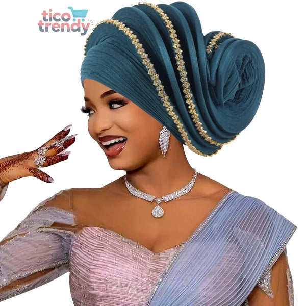 Mujer vistiendo un elaborando turbante azul petróleo estilo Gele con ribetes brillantes y dorados, combinado con un vestido formal de gala en tonos celestes y plateados.