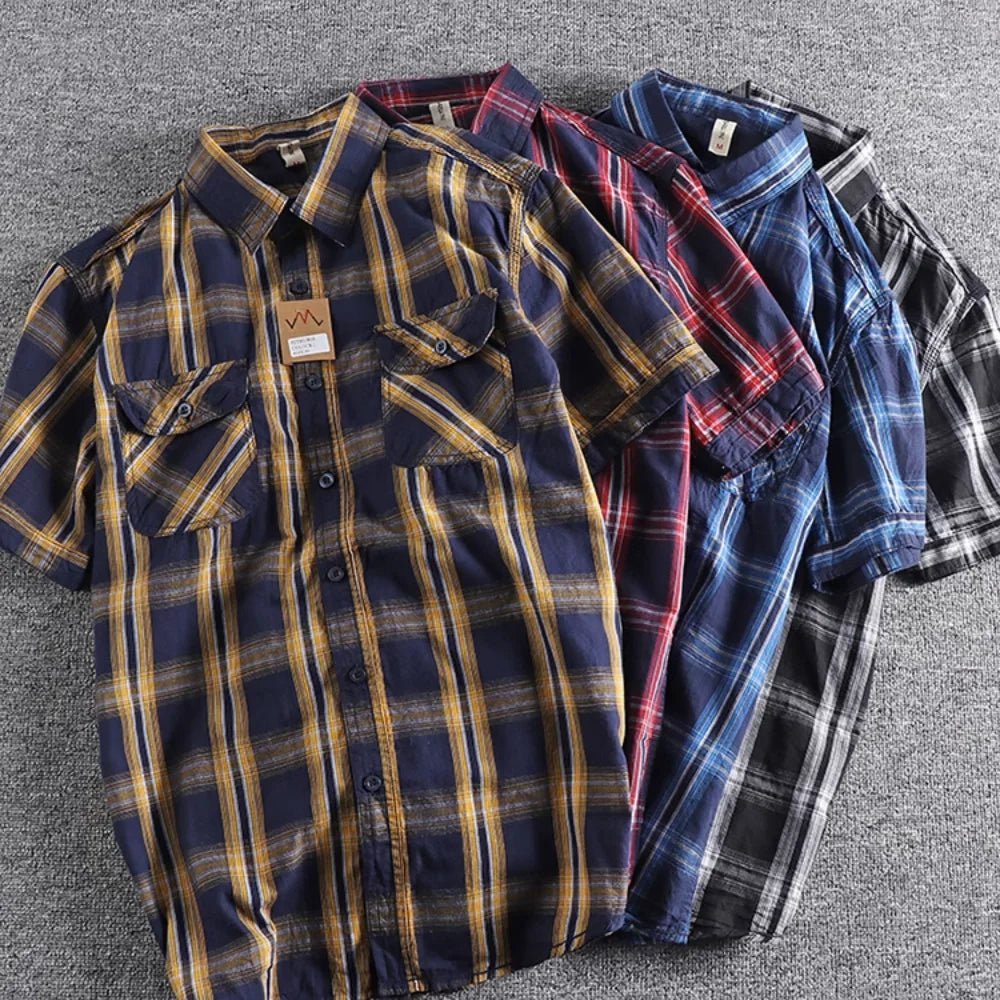 Cuatro camisas casuales de hombre de manga corta, con botones y bolsillos dobles en el pecho, apiladas y desplegadas ligeramente sobre una alfombra gris. incluyen: azul marino y amarillo/mostaza, rojo y azul, azul y blanco, y blanco y negro (gris).