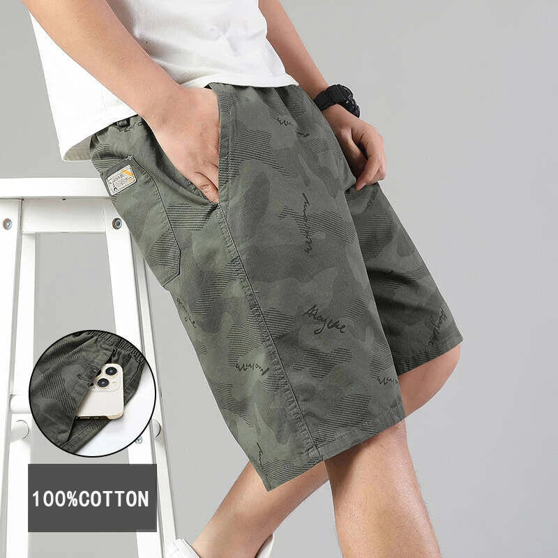 Plano medio de un hombre de pie, vistiendo una camiseta blanca y pantalones cortos cargo de estilo casual en color verde militar con un sutil estampado camuflado que incluye texto.