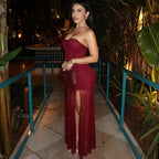Mujer vistiendo un vestido de fiesta largo, tubo, color burdeos o vino, con escote corazón strapless y una capa exterior de tul transparente con abertura lateral. Sostiene un bolso de mano a juego.