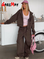 Mujer con cabello rubio en sweatsuit marrón y zapatillas de lona, posando junto a un mostrador en un ambiente casual. Muestra un look de moda urbana.