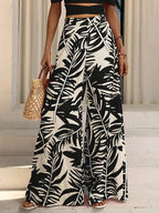 Mujer con crop top negro y pantalones anchos de pierna abierta con estampado de hoja de palma, sosteniendo un bolso de paja.