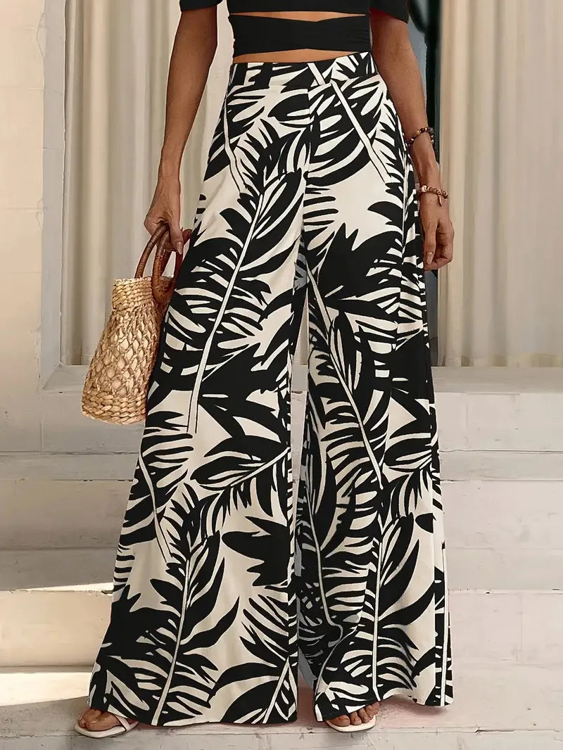 Mujer con crop top negro y pantalones anchos de pierna abierta con estampado de hoja de palma, sosteniendo un bolso de paja.