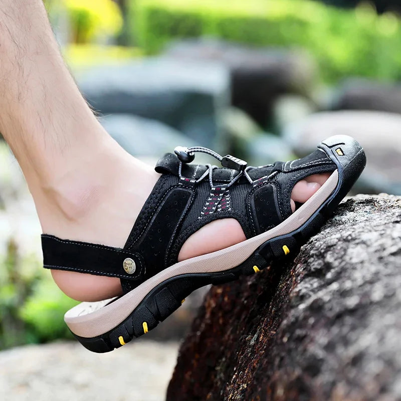 Primer plano de sandalias tipo trekking negras con sistema de cordones elásticos y suela gruesa, usadas por un hombre escalando una roca al aire libre.